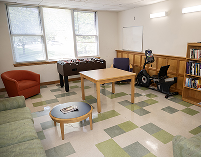 photo of lounge with two couches, two armchairs, a table, a coffee table, a foosball table, a bookcase with books, and a rowing machine