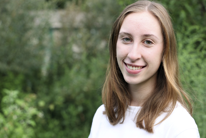 Emily, smiling and wearing a white shirt with the Campus Garden behind her. 